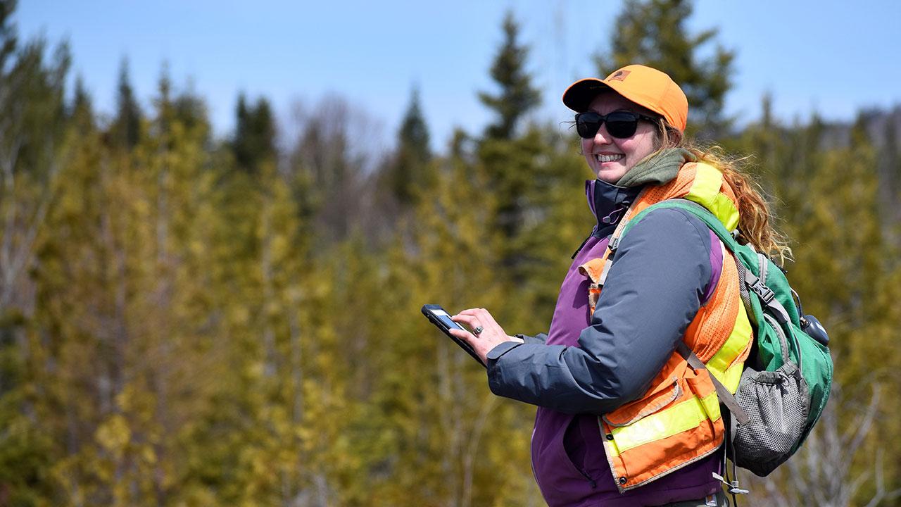 Tetra Tech employee with long hair in orange hat and vest types on a mobile device during a wind project field survey
