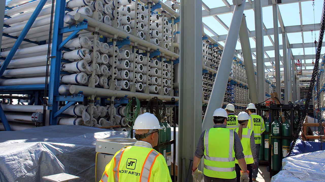 Tetra Tech employees wearing lime safety vests and hard hats walking in a line next to a stack of pipes